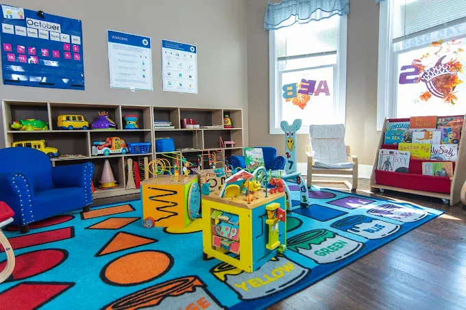 A bright playroom with a colorful shape rug, low toy shelves, a book display, and small reading chairs.