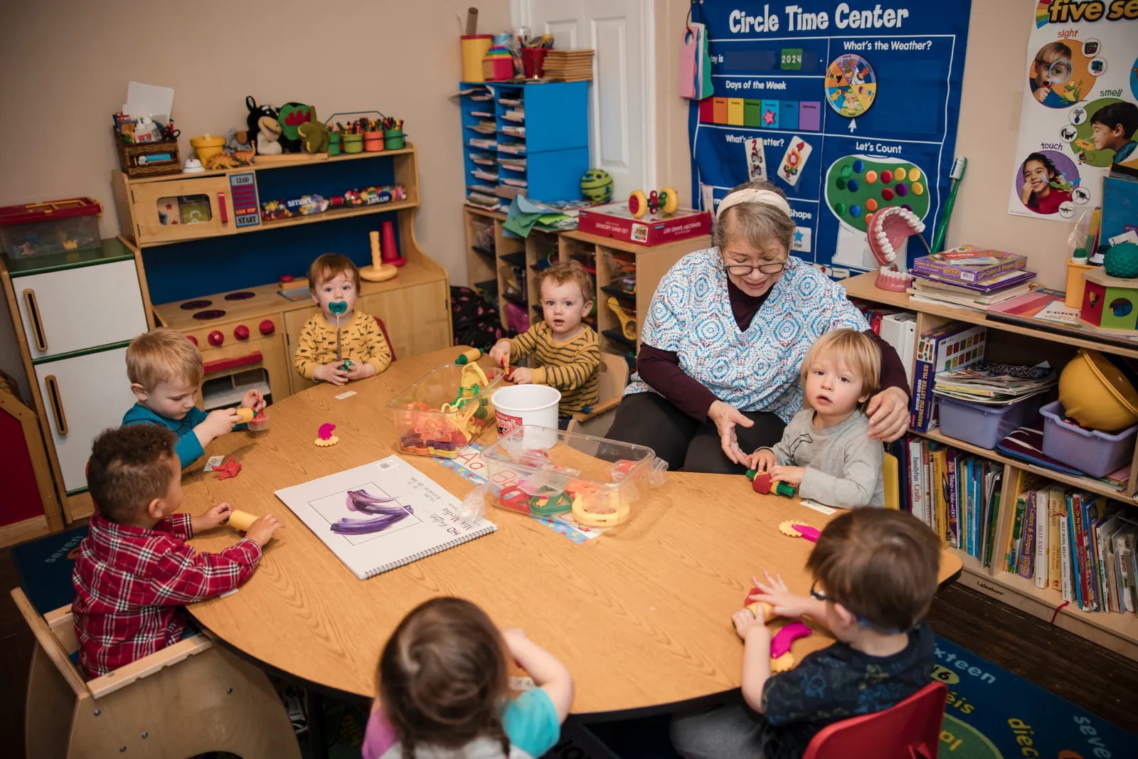 A caregiver and a group of children shaping playdough together around a round wooden table.
