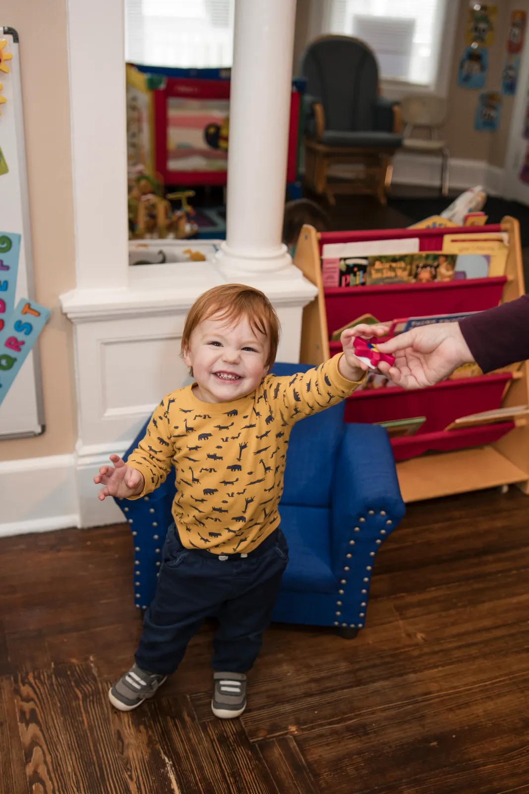 A smiling toddler standing by a small blue armchair, reaching for a caregiver's hand.