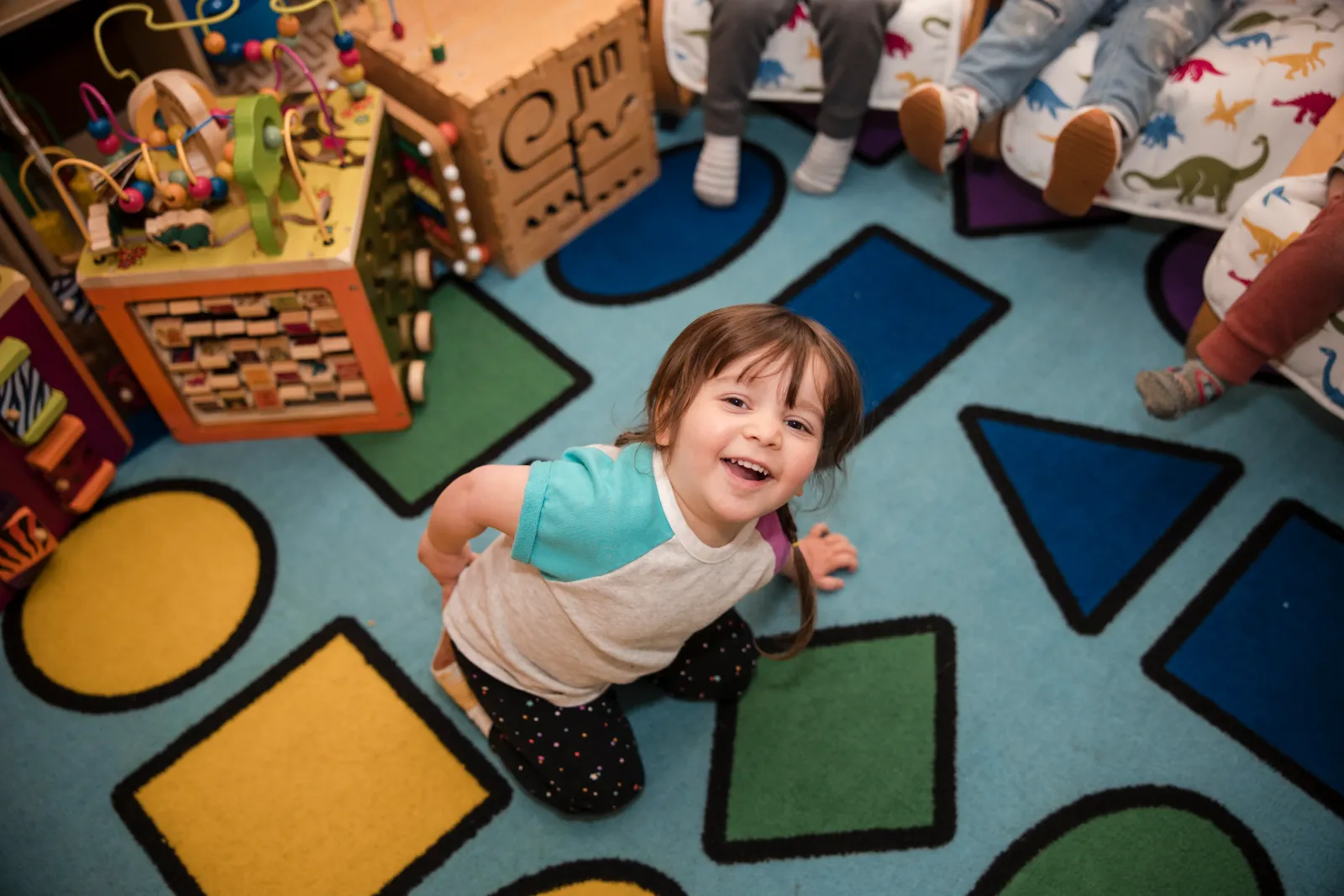 A child kneeling on the colorful shape rug, looking up with a big smile.
