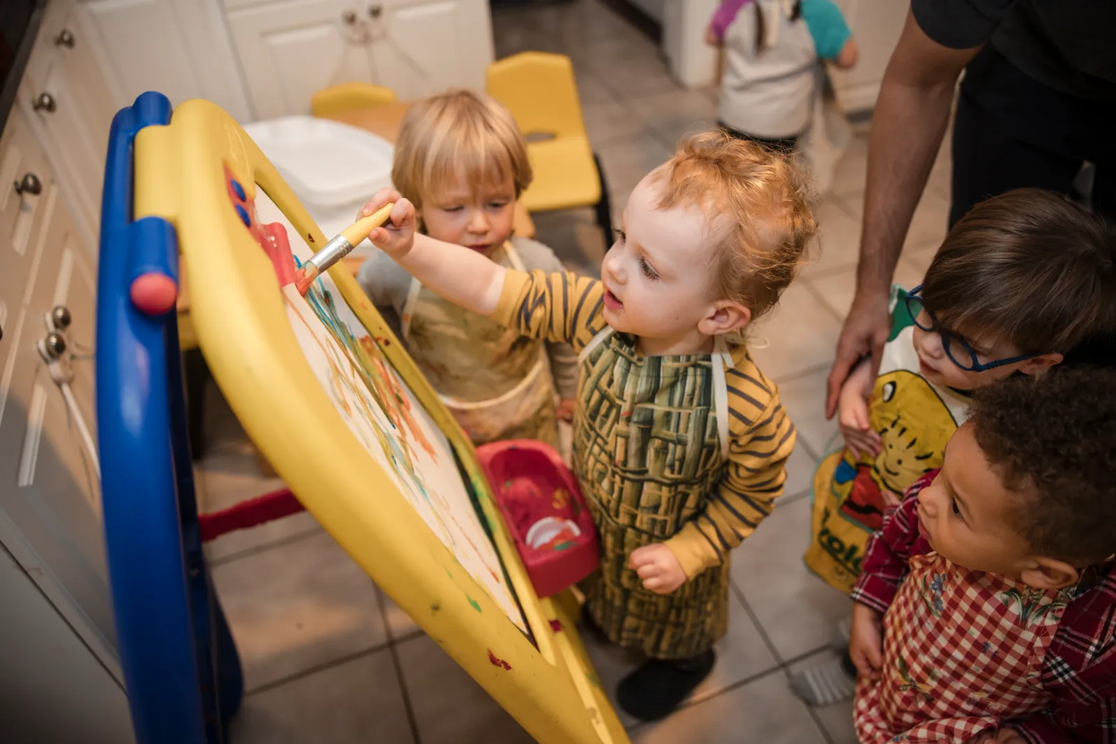 Two children painting at an easel while a caregiver helps nearby.