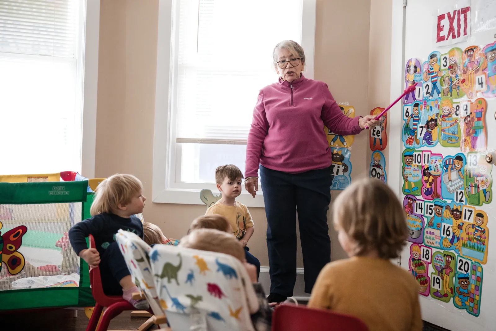 A caregiver points to a numbers chart on the wall as children look on.