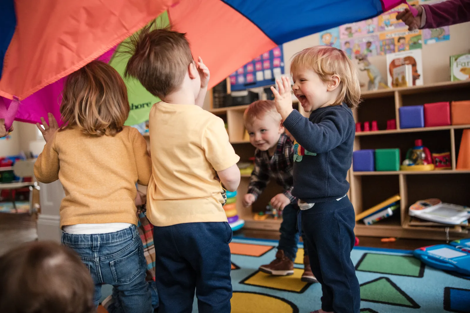 Children laughing as they lift a rainbow play parachute together with a caregiver.